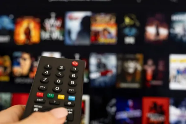 A hand holds a black TV remote control in the foreground, pointing towards a television screen that displays a blurred grid of movie and show thumbnails from a streaming service.